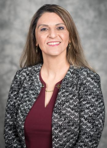 A woman with long, light brown hair, wearing a dark red blouse and great jacket smiles warmly at the viewer.