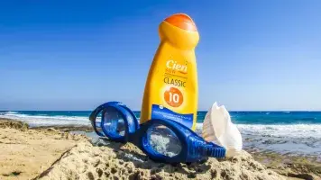 Bottle of sunscreen, goggles, and seashell sitting in the sand next to the ocean
